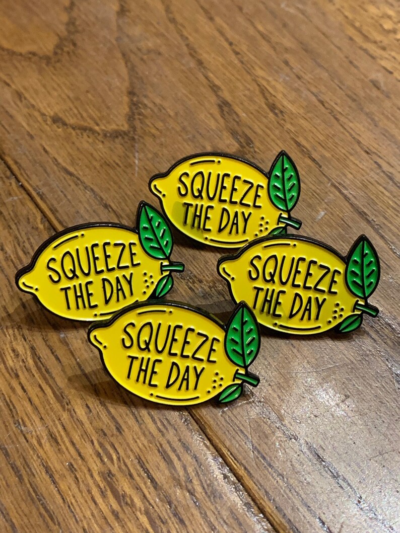 May include: Four yellow lemon-shaped enamel pins with black outlines and green leaves. Each pin says "Squeeze the Day" in black lettering.