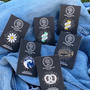 May include: A collection of eight enamel pins on black cards with white text. The pins feature a daisy, a wave with the text "Make Some Waves", a pretzel, a bus in a bottle, and two versions of the text "GRL PWR" on a banner. All cards have a cactus logo and the text "Crafty Cactus Company" and "Designed in Yorkshire".