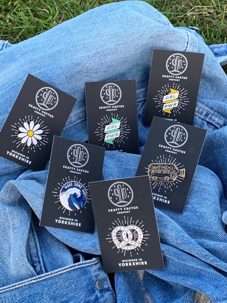 May include: A collection of eight enamel pins on black cards with white text. The pins feature a daisy, a wave with the text "Make Some Waves", a pretzel, a bus in a bottle, and two versions of the text "GRL PWR" on a banner. All cards have a cactus logo and the text "Crafty Cactus Company" and "Designed in Yorkshire".