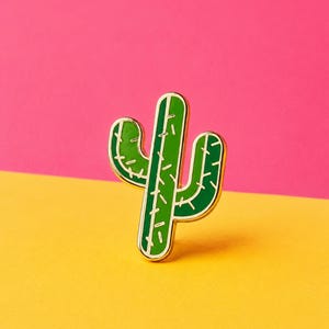 May include: A cactus-shaped enamel pin with gold-toned metal edges. The design features two shades of green enamel, with a lighter green on the central column and a darker green on the two side arms, accented by small, thin, gold-coloured lines.