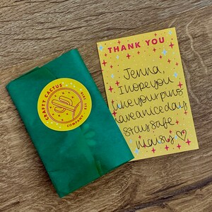 May include: A yellow thank you card with a handwritten message that says "Jenna, I hope you like your pins have a nice day Stay safe maisy". The card has a white background with blue and red stars. The card is sitting on a wooden surface.