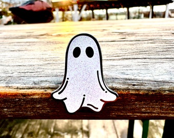 Puede incluir: Un pin de fantasma blanco con purpurina y ojos negros. El pin está sobre una superficie de madera.