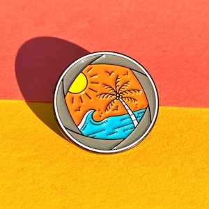 May include: A gray enamel pin with a camera lens design. Inside the lens is a cartoon image of a palm tree, sun, and ocean waves. The colors are orange, yellow, blue, and white.