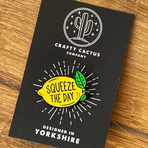 May include: A yellow enamel pin shaped like a lemon with green leaves. The pin says "Squeeze the Day" in black text. The pin is on a black background with a white cactus logo and the text "Crafty Cactus Company" and "Designed in Yorkshire".