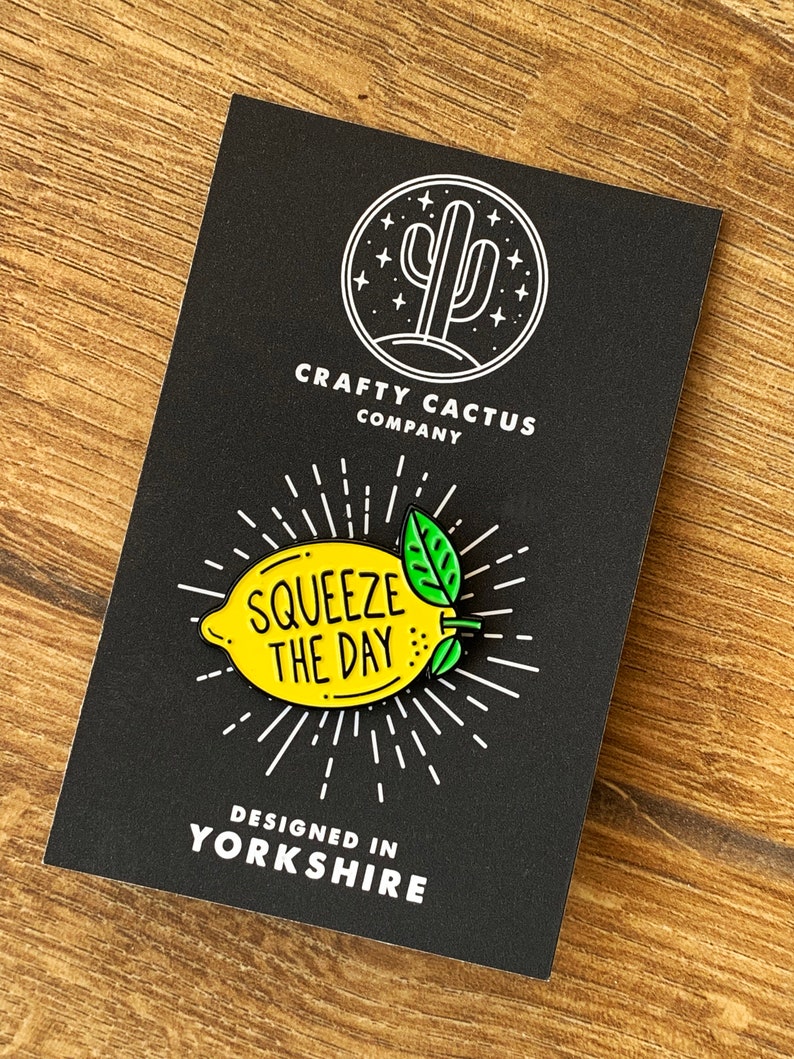 May include: A yellow enamel pin shaped like a lemon with green leaves. The pin says "Squeeze the Day" in black text. The pin is on a black background with a white cactus logo and the text "Crafty Cactus Company" and "Designed in Yorkshire".