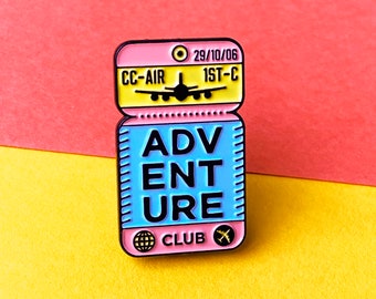 May include: A pink and blue enamel pin shaped like a boarding pass with the text "Adventure Club" and a small airplane icon. The top of the pin has the text "CC-AIR 29/10/06 1ST-C".