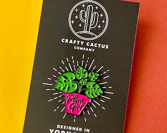 Puede incluir: Un pin de esmalte metálico que muestra una planta monstera verde en una maceta rosa con el texto Plant Girl, sobre una tarjeta negra con el texto Crafty Cactus Company y Designed in Yorkshire, rodeado de líneas blancas radiantes.