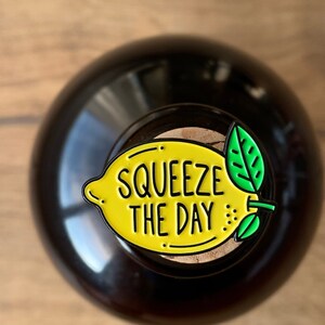 May include: A yellow lemon-shaped enamel pin with a green leaf and the text "Squeeze the Day" in black.