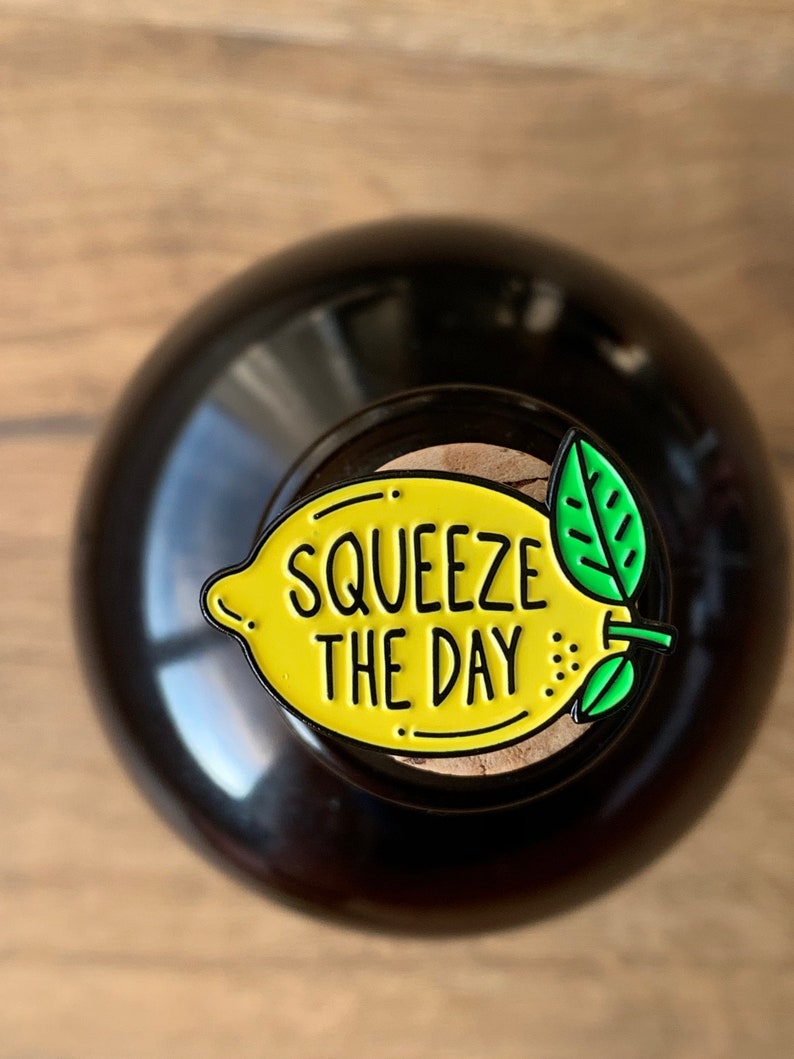 May include: A yellow lemon-shaped enamel pin with a green leaf and the text "Squeeze the Day" in black.