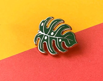 May include: A metal enamel pin shaped like a monstera leaf. The leaf is dark green with gold-colored metal outlines and thin white lines detailing the veins and fenestrations. The design features small white dots along the central vein area.