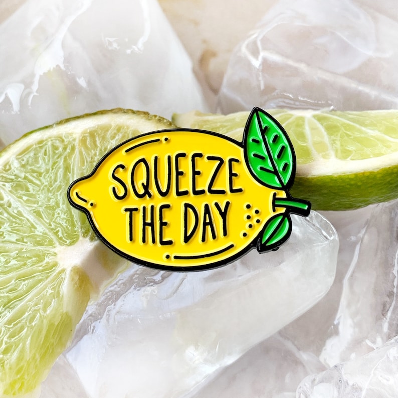 May include: A yellow enamel pin shaped like a lemon with the text "Squeeze the Day" in black. The pin is on top of a lime wedge and ice.