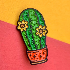 Op de afbeelding: Een groene cactus met gele bloemen in een rood-oranje pot. De cactus is zwart omlijnd en heeft een getextureerd oppervlak. De pot heeft een wit sterrenpatroon.