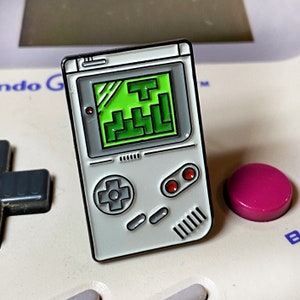 May include: A rectangular enamel pin shaped like a handheld video game console. The pin is light grey with a bright green screen displaying a block-stacking puzzle game. It features a directional pad, two red circular buttons, and two small oval buttons.