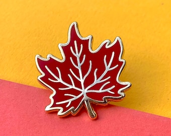 May include: A maple leaf enamel pin featuring a deep red fill with silver-toned metal outlines and vein details. The design captures the classic five-pointed shape of a maple leaf with a short stem at the bottom, finished with a polished metallic edge.