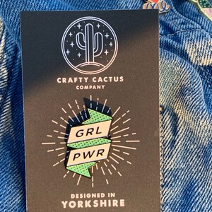 May include: A black and white illustration of a cactus with stars around it. The text "CRAFTY CACTUS COMPANY" is below the cactus. A green and white enamel pin with the text "GRL PWR" is shown on a black background. The text "DESIGNED IN YORKSHIRE" is below the pin.
