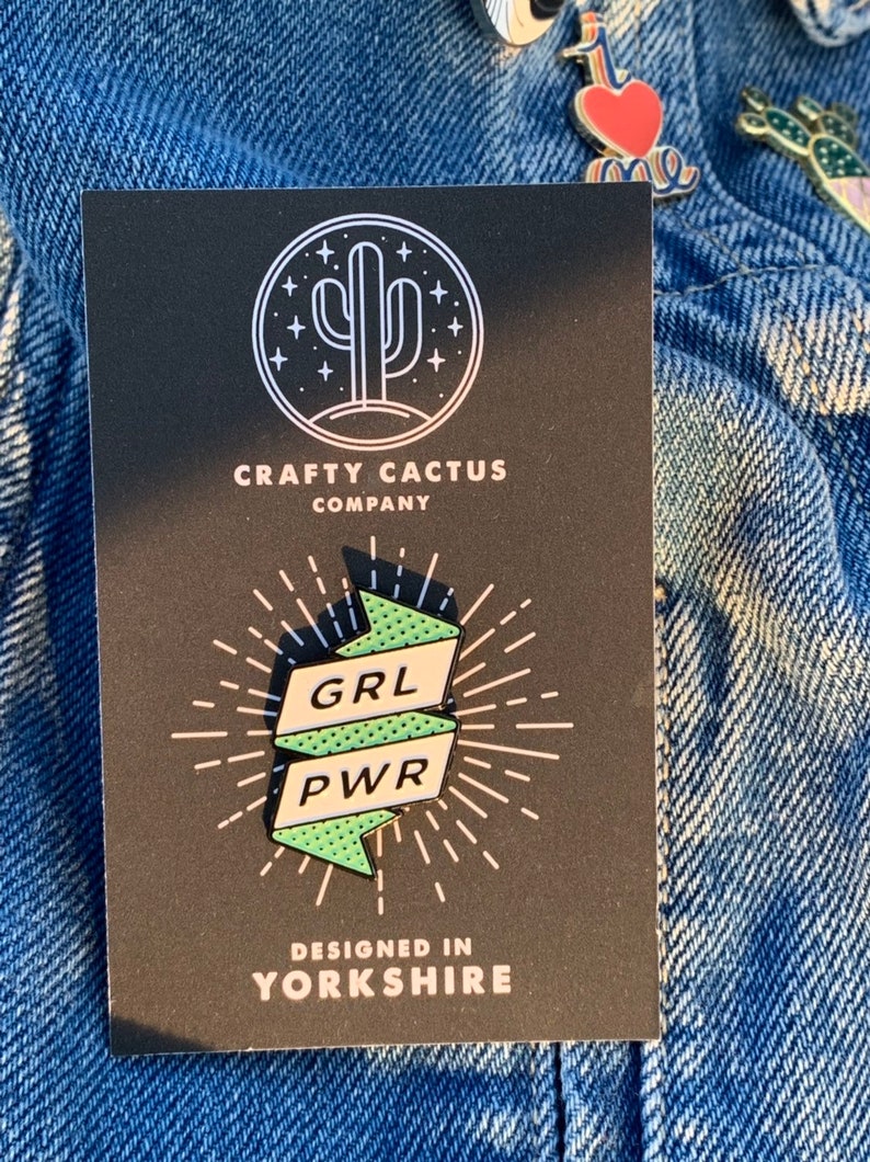 May include: A black and white illustration of a cactus with stars around it. The text "CRAFTY CACTUS COMPANY" is below the cactus. A green and white enamel pin with the text "GRL PWR" is shown on a black background. The text "DESIGNED IN YORKSHIRE" is below the pin.