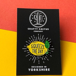 May include: A yellow enamel pin shaped like a lemon with green leaves and the text "Squeeze the Day" on it. The pin is on a black card with the text "Crafty Cactus Company" and "Designed in Yorkshire" on it.