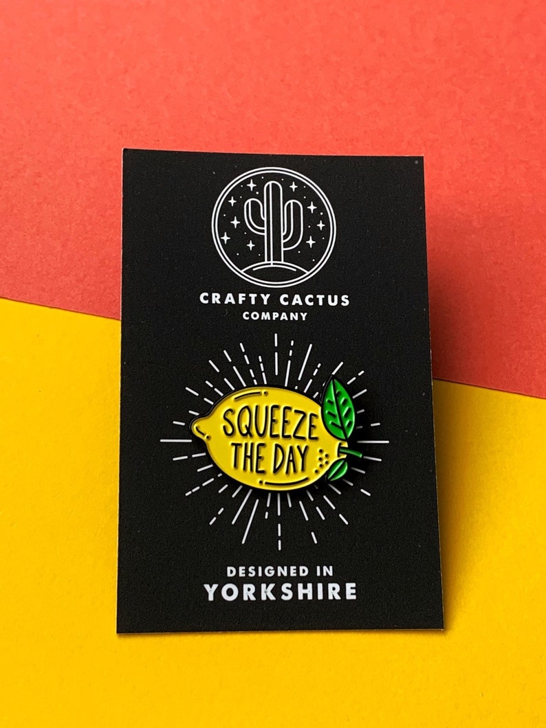 May include: A yellow enamel pin shaped like a lemon with green leaves and the text "Squeeze the Day" on it. The pin is on a black card with the text "Crafty Cactus Company" and "Designed in Yorkshire" on it.