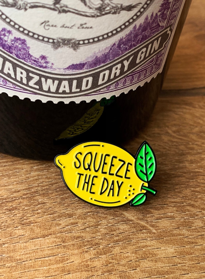 May include: Yellow enamel pin shaped like a lemon with the text "Squeeze the Day" in black. The pin has a green leaf on the stem.