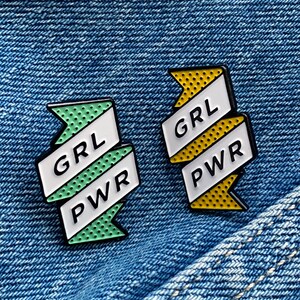 May include: Two enamel pins with a white ribbon design. The pins are on a blue denim background. The left pin is green with white polka dots and says "GRL PWR". The right pin is yellow with white polka dots and says "GRL PWR".