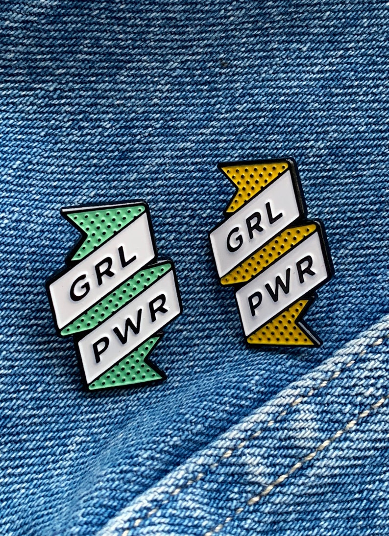May include: Two enamel pins with a white ribbon design. The pins are on a blue denim background. The left pin is green with white polka dots and says "GRL PWR". The right pin is yellow with white polka dots and says "GRL PWR".