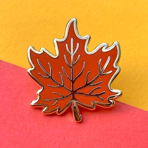May include: A gold-coloured enamel pin shaped like a maple leaf. The leaf is orange with black veins and a gold outline.