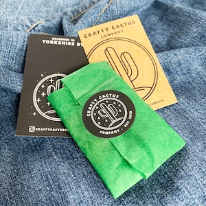 May include: A green tissue paper package with a black and white sticker that says "CRAFTY CACTUS COMPANY EST 2018" with a cactus graphic. The sticker is on a green tissue paper package. The package is on top of a blue denim fabric.