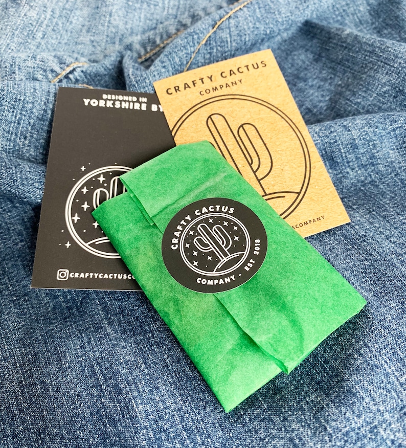 May include: A green tissue paper package with a black and white sticker that says "CRAFTY CACTUS COMPANY EST 2018" with a cactus graphic. The sticker is on a green tissue paper package. The package is on top of a blue denim fabric.