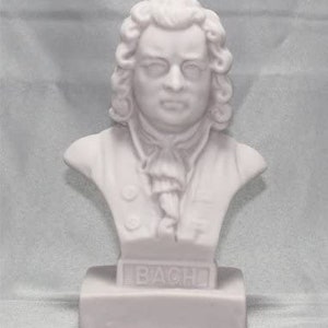 May include: White plaster bust of Johann Sebastian Bach, a famous composer. The bust is on a white base with the word "BACH" inscribed on it.