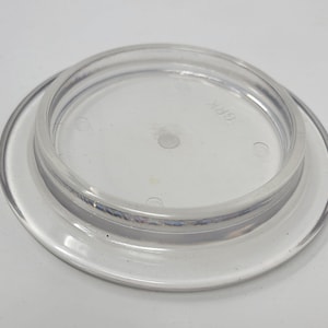 May include: A transparent plastic lid, circular in shape, with a raised inner ring. The lid's clear design allows visibility of the surface below. It features a slightly recessed centre and a smooth, rounded edge. The text "GRK" is visible on the lid.