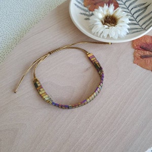 Peut inclure: Un bracelet en perles dorées et irisées avec une fermeture en cordon marron. Le bracelet est posé sur une surface en bois brun clair.