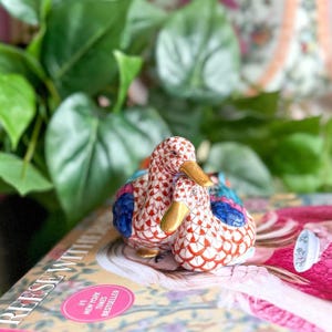 May include: Two ceramic duck figurines. The ducks are white with red and blue patterned details, gold beaks, and are nestled together. The figurines are on a book with the text "#1 New York Times Bestseller".