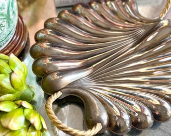 May include: A silver serving tray shaped like a seashell with two rope handles. The tray has a shiny, metallic finish and a fan-like design.
