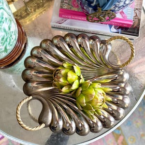 May include: A silver serving tray with a scalloped edge and two gold handles. The tray is filled with two artichoke hearts.