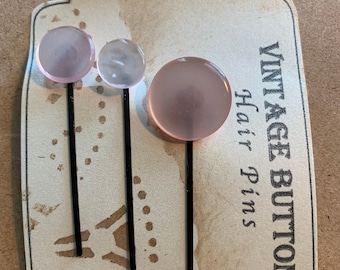 Könnte beinhalten: Drei Vintage-Haarnadeln mit rosa, runden Knöpfen und schwarzen Stielen. Die Haarnadeln werden auf einem Etikett im Vintage-Stil mit der Aufschrift "VINTAGE BUTTON Hair Pins" und der Nummer "72" präsentiert.