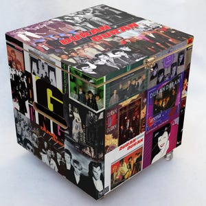 Pode incluir: Uma caixa de armazenamento quadrada coberta com arte de álbuns e fotos da banda Duran Duran. A caixa apresenta uma tampa articulada com um fecho de latão e pequenos pés prateados. A caixa é uma colagem de capas de álbuns e imagens da banda.