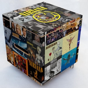 Pode incluir: Uma caixa de armazenamento de madeira com uma colagem de arte de álbuns do Nirvana e imagens da banda. A caixa tem uma tampa articulada e uma alça recortada. A parte superior da caixa apresenta o logotipo do Nirvana e um smiley amarelo. A caixa fica sobre pequenas rodas prateadas.