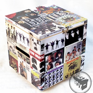 Könnte beinhalten: Eine Holzkiste, die mit Vintage-Beatles-Albumcovern bedeckt ist. Die Kiste hat einen Klappdeckel und einen Metallverschluss. Die Kiste ist mit verschiedenen Albumcovern verziert, darunter "Sgt. Pepper's Lonely Hearts Club Band", "Abbey Road" und "Rubber Soul".