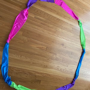 May include: A colorful fabric streamer with six sections in pink, purple, green, blue, purple, and pink. The streamer is tied in a circle and laid out on a wooden floor.
