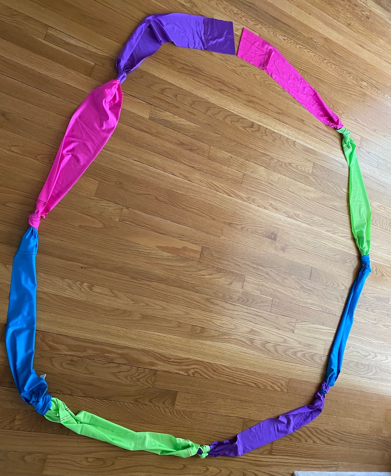 May include: A colorful fabric streamer with six sections in pink, purple, green, blue, purple, and pink. The streamer is tied in a circle and laid out on a wooden floor.