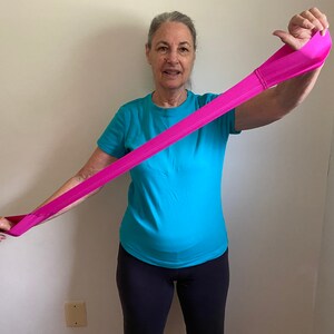 May include: A person demonstrates a pink resistance band exercise by stretching the band overhead with one arm extended.