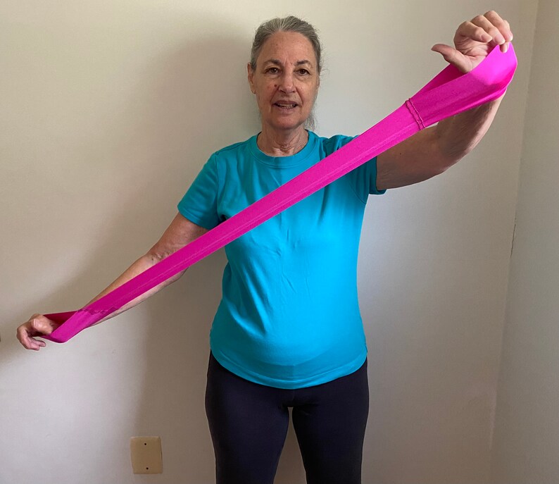 May include: A person demonstrates a pink resistance band exercise by stretching the band overhead with one arm extended.