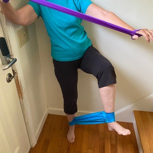 May include: A person is doing a leg exercise using a purple resistance band. The band is looped around their ankle and they are holding the other end of the band with their hand. They are standing on one leg and lifting the other leg up.