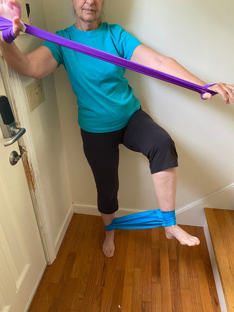 May include: A person is doing a leg exercise using a purple resistance band. The band is looped around their ankle and they are holding the other end of the band with their hand. They are standing on one leg and lifting the other leg up.