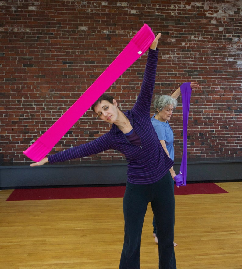 May include: Two people are stretching with pink and purple resistance bands. The woman in the foreground is holding a pink resistance band above her head and stretching her arm to the side. The woman in the background is holding a purple resistance band and stretching her arm to the side.