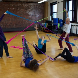 May include: Four people are using colorful fabric strips to create a geometric shape in a dance studio. The strips are pink, blue, purple, and green. The people are all wearing casual clothing and are smiling.