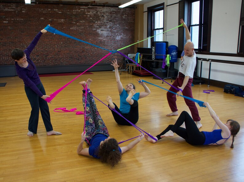 May include: Four people are using colorful fabric strips to create a geometric shape in a dance studio. The strips are pink, blue, purple, and green. The people are all wearing casual clothing and are smiling.