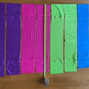 May include: Six strips of fabric in different colors: purple, pink, green, and blue. The fabric strips are laid out on a wooden surface with a measuring tape next to them.