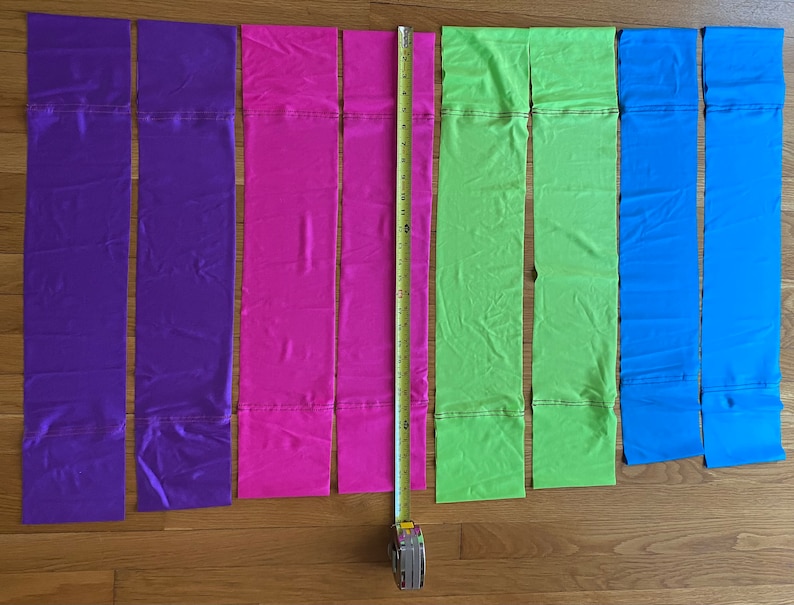 May include: Six strips of fabric in different colors: purple, pink, green, and blue. The fabric strips are laid out on a wooden surface with a measuring tape next to them.