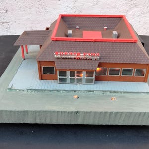 May include: Model Burger King restaurant with brown roof tiles, red trim, and the Burger King logo. The building has windows and lights, and sits on a grey base. A detailed replica of the fast-food restaurant.